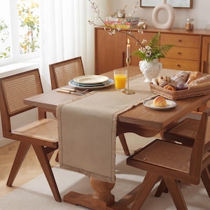 Op de afbeelding: Een rechthoekige beige tafelloper met gerafelde randen op een houten eettafel. De tafel is omringd door vier houten stoelen met zittingen en rugleuningen van rietvlechtwerk, gedekt met borden, een glas sinaasappelsap en een broodmandje.