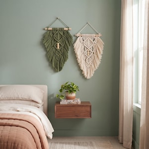 Può includere: Due arazzi in macramè appesi a bastoni di legno, uno verde oliva e uno color crema. Ogni pezzo presenta intricati motivi a nodo, bordi sfrangiati e tre perline di legno centrali, creando una forma simmetrica simile a una foglia.