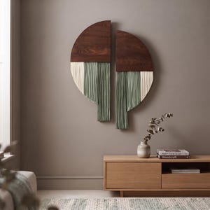 Op de afbeelding: Een tweedelige wandhanger met donkerbruine houten halve cirkels aan de bovenkant en lange, verticale vezelfranjes in afwisselende tinten crème en saliegroen die in een gelaagde, asymmetrische opstelling naar beneden hangen.