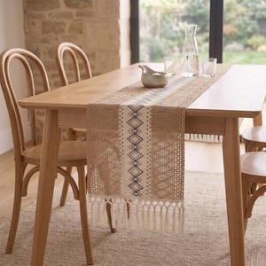 May include: A rectangular table runner featuring a central cream-colored panel with a blue and brown geometric diamond pattern, flanked by textured tan woven sections and finished with long, cream-colored fringe tassels at both ends.