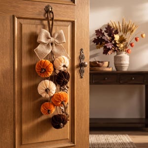 May include: A hanging door decoration featuring a large tan burlap bow at the top, with several small velvet pumpkins in shades of orange, cream, and dark brown suspended below. The pumpkins are arranged in a cluster on a vertical stem structure.