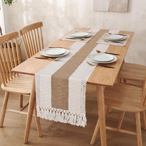 May include: A rectangular table runner featuring a central strip of natural tan burlap flanked by white crocheted lace panels with intricate patterns. The runner ends in long, white fringe tassels and is set on a light wood dining table.