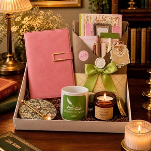May include: A gift box containing a pink notebook with a gold clasp, a brown envelope with a green ribbon, a white mug with a green graphic and the text "BETWEEN THE BOOK LINES", a floral coaster with a gold spoon, and a rosemary scented candle.