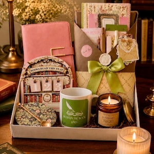 May include: A gift box containing a pink journal, a "Books Read This Year" tracker, a white mug with a green "Between the Book Lines" graphic, a rosemary candle, a floral coaster with a gold spoon, and a brown envelope tied with a green ribbon.
