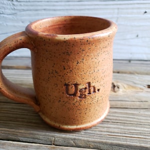 May include: A speckled brown ceramic mug with a handle and the word "Ugh." inscribed on the front.