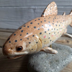 May include: A ceramic sculpture of a brown trout with black spots and pink markings. The fish is mounted on a gray stone base.