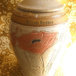 May include: A ceramic urn with a lid, featuring a beige and brown color scheme. The urn is adorned with a poppy flower design in shades of peach and brown. The name "Kristi Foster" is inscribed on the urn.