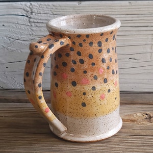 May include: A ceramic mug with a speckled brown, yellow, and white glaze. The mug has a large handle with black and pink accents. The mug is decorated with black and pink dots.