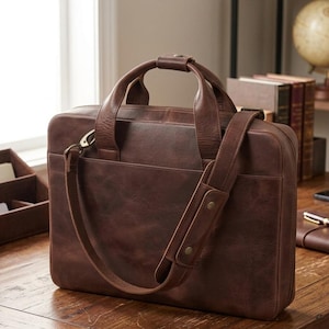 May include: A dark brown leather briefcase featuring a rectangular silhouette, two top handles, and a detachable shoulder strap with a padded section. The bag includes a front exterior pocket, metal hardware, and a smooth, matte finish throughout.