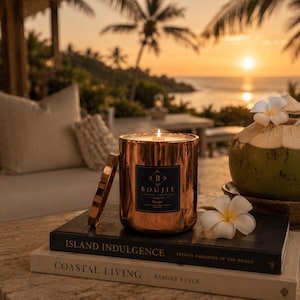 May include: A lit candle in a metallic copper-colored cylindrical vessel with a black label reading Boujie Candle Company, placed on two stacked books titled Island Indulgence and Coastal Living, next to a fresh coconut with a white flower.