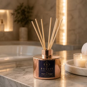 May include: A copper-colored cylindrical reed diffuser with a dark blue label featuring the text Boujie Candle Company and Boujie on a Budget. The diffuser has a metallic finish and contains several light-colored wooden reeds extending upwards.