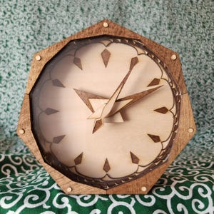 Peut inclure: Une horloge murale hexagonale en bois avec un cadran clair orné de marqueurs d'heures géométriques marron foncé et d'aiguilles pointues assorties. Le cadre est en bois teinté au grain visible, et le cadran est protégé par une vitre.