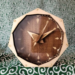 May include: A hexagonal wall clock featuring a light-coloured wooden frame and a dark brown wooden face. The clock has sharp, angular wooden hands and a series of small, diamond-shaped light wood markers arranged in a circular pattern on the face.