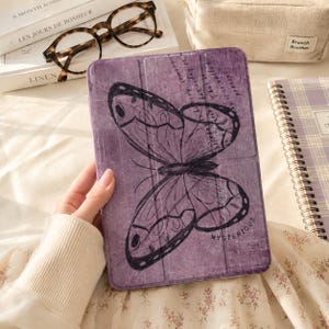 May include: A purple tablet case featuring a large, black, hand-drawn style butterfly illustration. The case has a textured, distressed surface finish and includes the word "MYSTERIOUS" printed in small black letters near the bottom right corner.