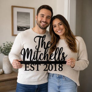 Może przedstawiać: Czarny, wycinany laserowo szyld z napisem "The Mitchells" wykonanym kursywą nad poziomą linią oraz napisem "EST 2018" pogrubioną czcionką bezszeryfową. Szyld wykonany jest z płaskiego materiału o matowym wykończeniu.