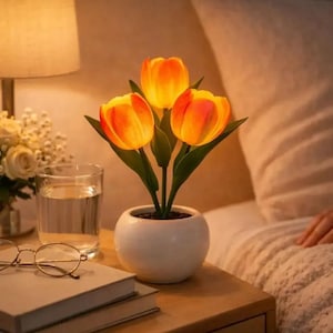 May include: A decorative lamp shaped like three orange tulips with green stems and leaves, potted in a small white ceramic bowl. The tulip petals emit a warm, soft glow, creating a gentle illumination against the surrounding room environment.
