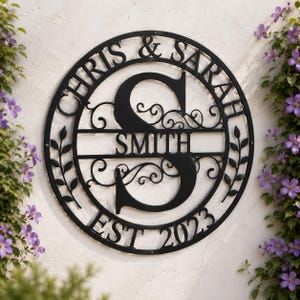 Puede incluir: Un letrero de pared circular de metal negro con una letra S grande en el centro, el nombre SMITH, el texto CHRIS & SARAH y la fecha EST 2023. El diseño incluye volutas decorativas y motivos de hojas dentro del marco circular.