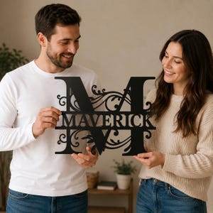 May include: A black metal wall sign featuring a large capital letter M with intricate scrollwork and flourishes. The name MAVERICK is cut out in capital letters across the centre of the letter M, creating a personalised decorative piece.
