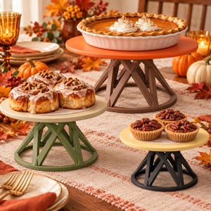 May include: Three tiered cake stands with geometric bases in orange, green, and black. The orange stand holds a pumpkin pie with whipped cream, the green stand holds frosted cinnamon rolls, and the black stand holds three pecan tarts.