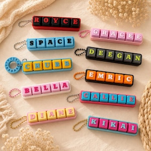 May include: A collection of rectangular keychains featuring mechanical keyboard-style keys with letters spelling names including ROYCE, SPACE, GRACEE, DEEGAN, CHUBB, EMRIC, BELLA, CALLIE, DIANE, and KIKAI, each attached to a small metal ball chain.