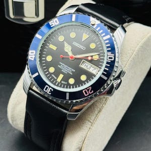 May include: A Seiko automatic wristwatch with a black dial, luminous circular hour markers, and a red second hand. The watch features a blue and black rotating bezel, a day-date window at the three o'clock position, and a black leather strap.