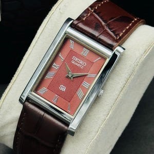 Könnte beinhalten: Eine rechteckige Seiko Quartz Armbanduhr mit silberfarbenem Gehäuse, einem strukturierten lachsfarbenen Zifferblatt mit silbernen römischen Ziffern als Stundenmarkierungen und einem dunkelbraunen Lederarmband mit Krokodilprägung.