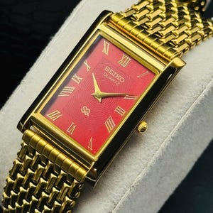 Könnte beinhalten: Eine rechteckige, goldfarbene Seiko Quartz Armbanduhr mit einem leuchtend roten, strukturierten Zifferblatt. Die Uhr verfügt über goldfarbene römische Ziffern als Stundenmarkierungen, goldfarbene Zeiger und ein passendes Metallgliederarmband.