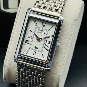 May include: A rectangular Seiko Quartz wristwatch with a white dial, black Roman numeral hour markers, and gold-tone hands. The watch features a silver-tone metal link bracelet with a textured, multi-row design and a small logo at the bottom.