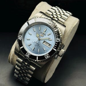 May include: A silver-tone Seiko 5 Sports automatic wristwatch featuring a light blue dial, a black rotating bezel with white numerals, and a stainless steel jubilee-style link bracelet. The dial displays the Seiko logo, day-date window, and markers.