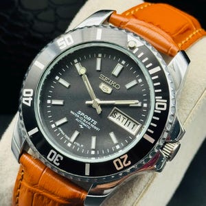 May include: A Seiko 5 Sports automatic wristwatch featuring a black dial with luminous markers, a day-date display, and a black rotating bezel with silver numerals. The watch has a stainless steel case and a textured orange leather strap.