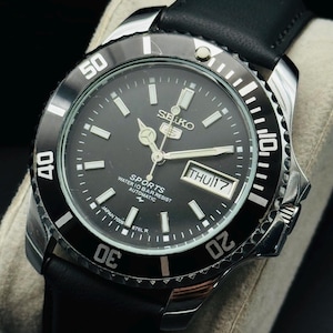 May include: A Seiko 5 Sports automatic wristwatch with a black dial, silver-tone hands and hour markers, and a black rotating bezel with white numerals. The watch features a day-date window at the three o'clock position and a black leather strap.