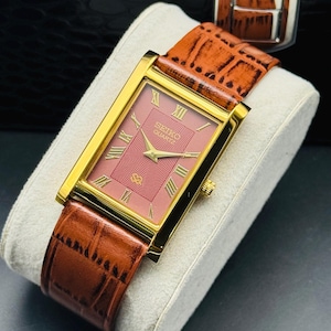 Könnte beinhalten: Eine rechteckige Seiko Quartz Armbanduhr mit goldfarbenem Gehäuse und einem strukturierten, altrosa Zifferblatt mit goldenen römischen Ziffern. Die Uhr ist mit einem braunen, krokodilgeprägten Lederarmband mit goldfarbener Schließe ausgestattet.