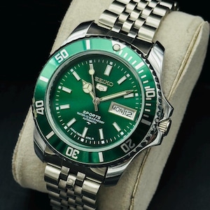 May include: A Seiko 5 Sports automatic wristwatch featuring a vibrant green dial and a matching green rotating bezel with white numerals. The watch has a stainless steel case and a multi-link metal bracelet, with a day-date window at the 3 o'clock position.