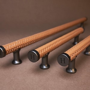 Peut inclure: Trois poignées de placard de longueurs variables dotées d'un revêtement en cuir tressé beige autour d'une barre cylindrique. Chaque poignée possède des supports de montage en métal foncé et des embouts circulaires avec la lettre H gravée.