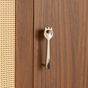 May include: A polished silver-toned metal cabinet handle shaped like a fork, mounted on a vertical wooden cabinet door with a visible wood grain pattern, positioned next to a section of light-colored woven cane webbing material.