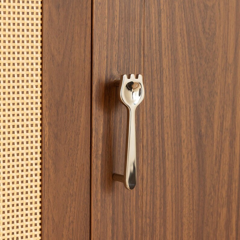 May include: A polished silver-toned metal cabinet handle shaped like a fork, mounted on a vertical wooden cabinet door with a visible wood grain pattern, positioned next to a section of light-colored woven cane webbing material.