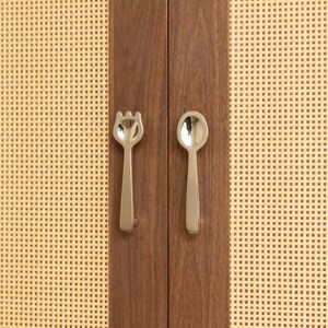 May include: A pair of silver-toned metal cabinet handles shaped like a fork and a spoon, mounted on a dark brown wooden cabinet door with light beige woven cane panels on either side of the vertical wooden frame holding the hardware.