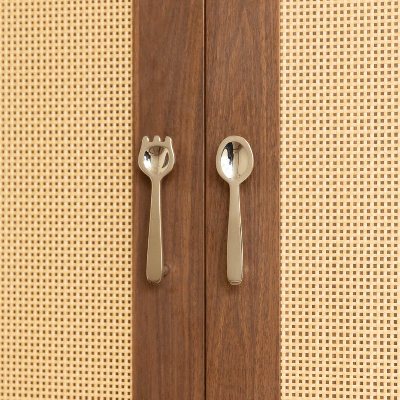 May include: A pair of silver-toned metal cabinet handles shaped like a fork and a spoon, mounted on a dark brown wooden cabinet door with light beige woven cane panels on either side of the vertical wooden frame holding the hardware.