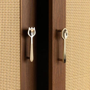 May include: Two silver-toned metal cabinet pulls shaped like a fork and a spoon, mounted on dark brown wooden cabinet doors with light beige woven cane panels. The fork-shaped handle is on the left, and the spoon-shaped handle is on the right.