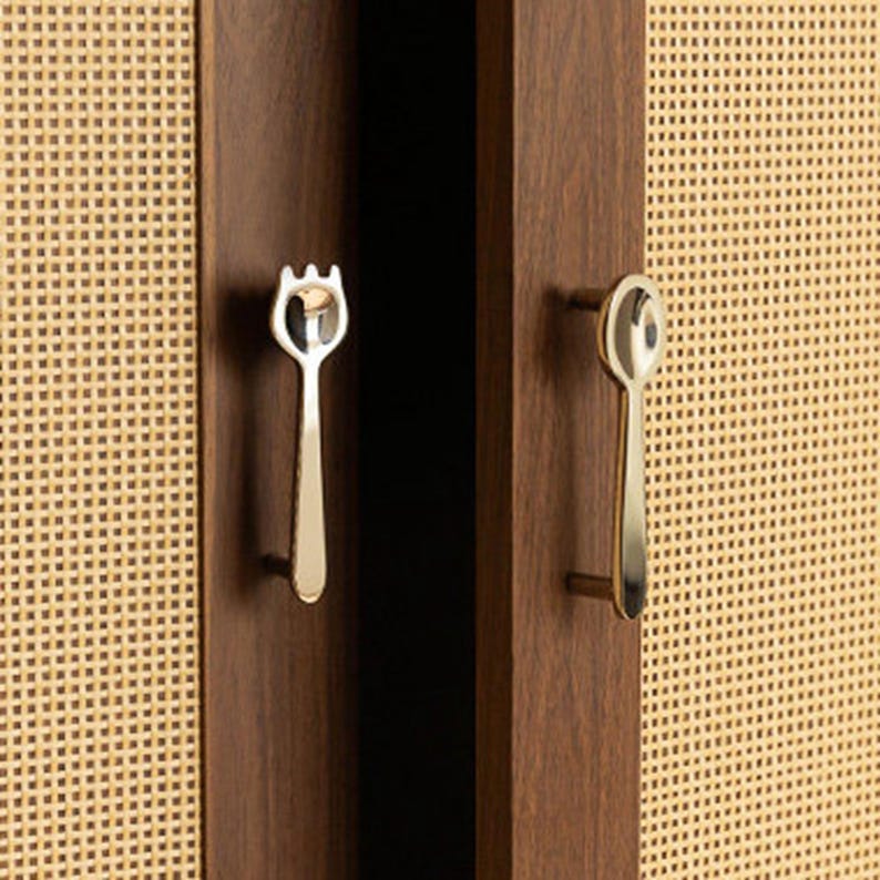 May include: Two silver-toned metal cabinet pulls shaped like a fork and a spoon, mounted on dark brown wooden cabinet doors with light beige woven cane panels. The fork-shaped handle is on the left, and the spoon-shaped handle is on the right.