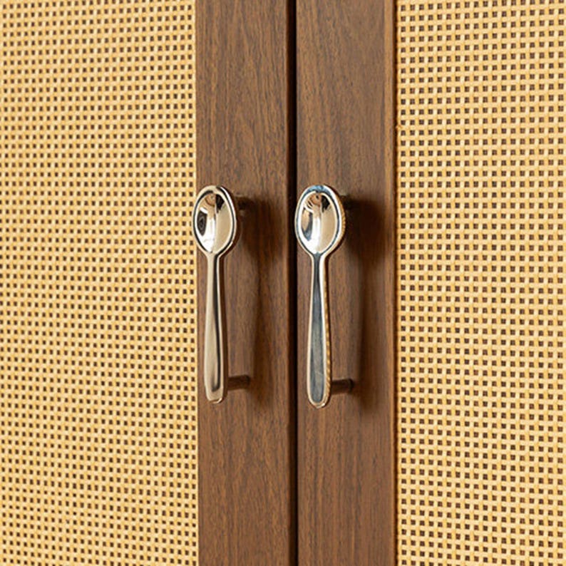 May include: Two silver-toned metal cabinet handles shaped like spoons attached to dark brown wooden cabinet doors with a light tan woven cane mesh paneling. The handles are vertically oriented and feature a polished, reflective metallic finish.
