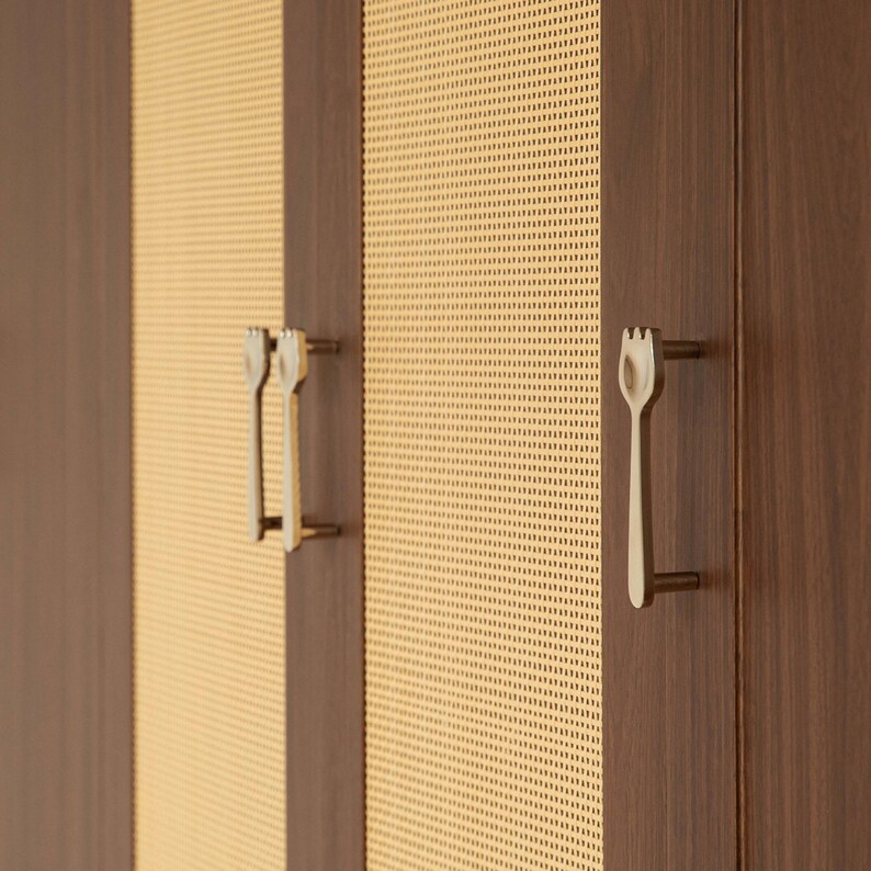 May include: A cabinet featuring dark brown wood panels alternating with light beige woven cane mesh sections. The doors are equipped with metallic, fork-shaped handles that have a distinct three-pronged top design and a vertical stem.