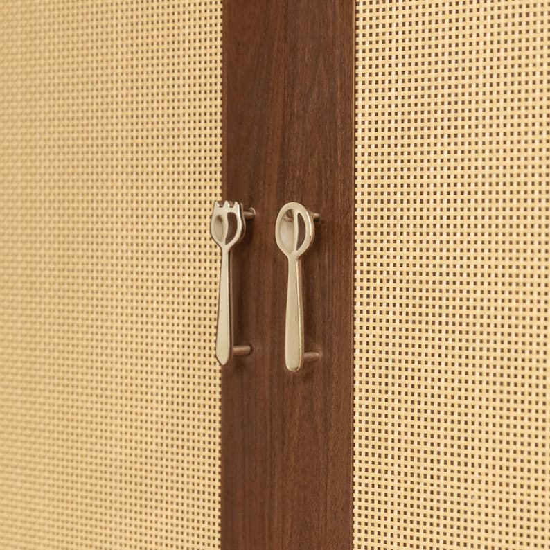 May include: A dark brown wooden cabinet door panel featuring a light beige woven cane texture on both sides. Two silver-toned metal cabinet pulls are mounted vertically, one shaped like a fork and the other shaped like a spoon, centered on the wood.