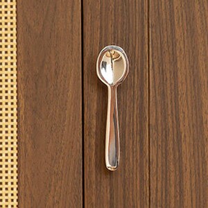 May include: A small, polished silver-toned metal spoon with a rounded bowl and a smooth, straight handle. The spoon features a reflective, metallic surface that catches light, creating bright highlights along its curved edges and center.