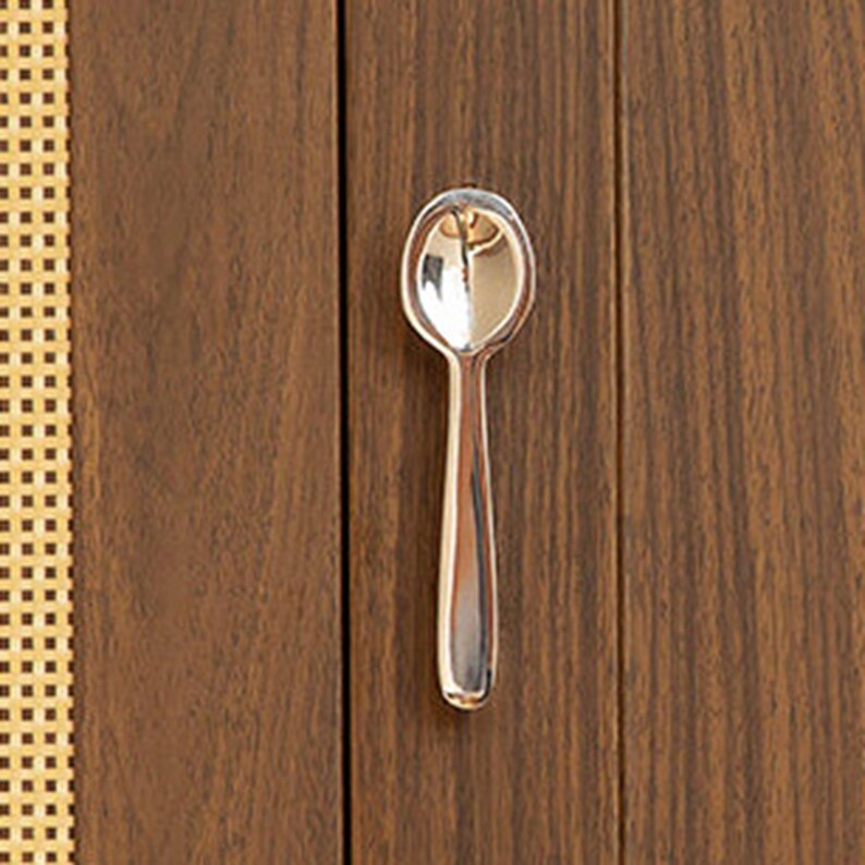 May include: A small, polished silver-toned metal spoon with a rounded bowl and a smooth, straight handle. The spoon features a reflective, metallic surface that catches light, creating bright highlights along its curved edges and center.
