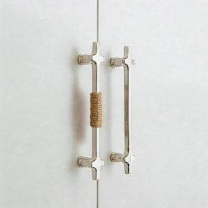 May include: Two vertical silver-toned metal cabinet pulls mounted on a white surface. The left pull features a tan jute rope wrapped around its centre. The right pull has the text CrimsonQuiltShop engraved vertically along its central bar.