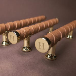 Peut inclure: Trois poignées de placard cylindriques dotées d'un revêtement en cuir texturé beige autour de la tige. Chaque poignée possède des embouts et des supports de fixation en métal doré poli. Les embouts circulaires portent la lettre H gravée.