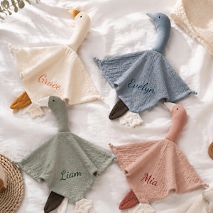 May include: Four soft muslin security blankets shaped like geese in cream, blue, sage green, and dusty pink. Each blanket features an embroidered name: Grace, Evelyn, Liam, and Mia. The blankets have long necks, small heads, and attached fabric feet.