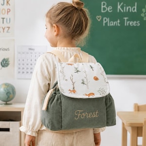 May include: A sage green corduroy backpack featuring a white flap with a forest-themed print of trees and foxes. The word Forest is embroidered in gold thread on the front pocket. The bag includes light beige straps and a top handle.
