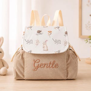 May include: A beige corduroy backpack featuring a white flap with a printed pattern of rabbits, snails, and botanical elements. The word Gentle is embroidered in brown thread on the front. The bag includes light beige straps and side pockets.